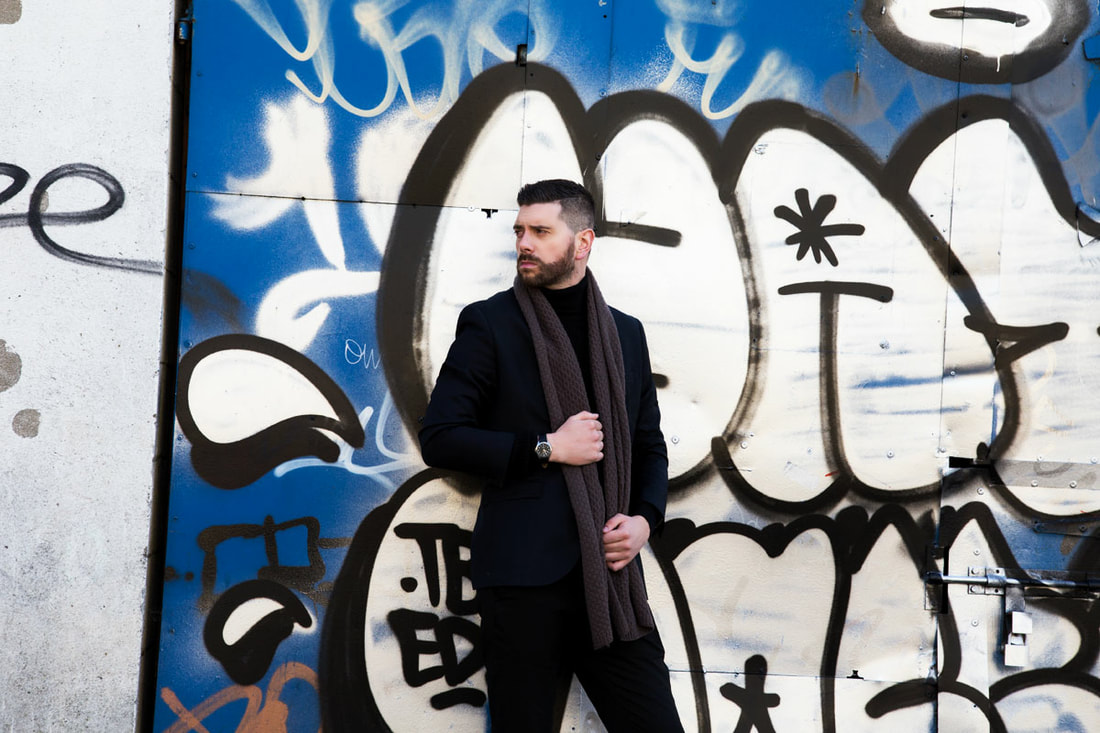 A male model with scarf posing in front of the wall with graffiti on it