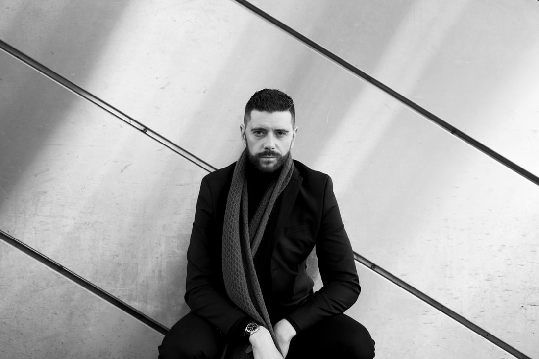 Male model with scarf, black high neck jumper and suit jacket, posing in front of a modern building