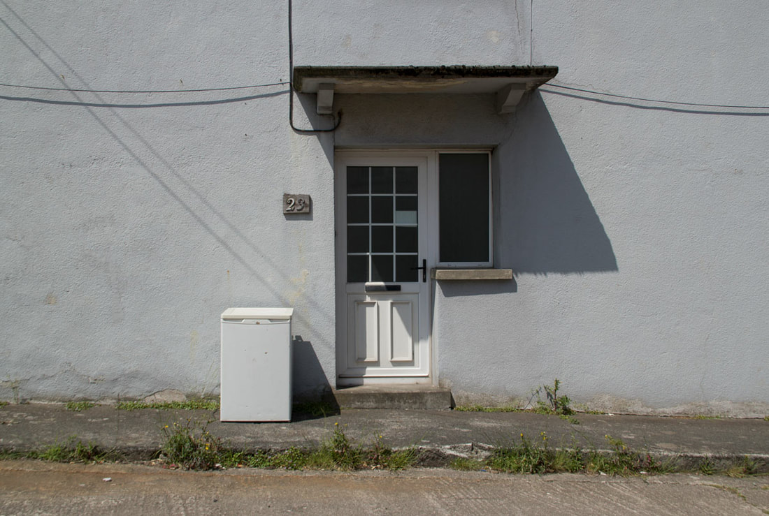 A grey wall with a white door and a small white fridge in front of it. 