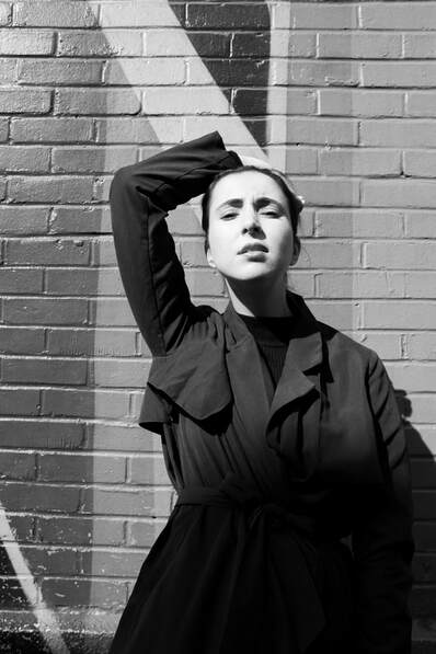 A girl with one arm over her head wearing a trench coat standing in front of a wall