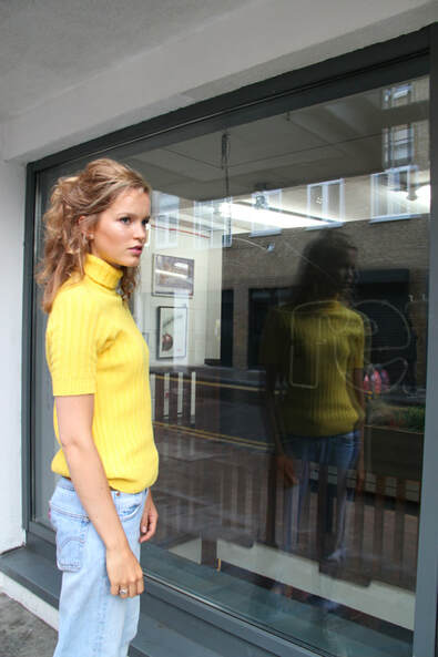 Female model with yellow outfit in front of glass with her reflection in it 