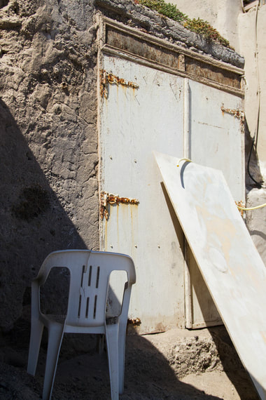 A rusty white door and a white chair on the beach with a shadow across them. 