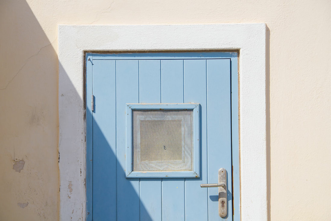 A shadow cuts through the light blue door with white frame on a yellow wall. 