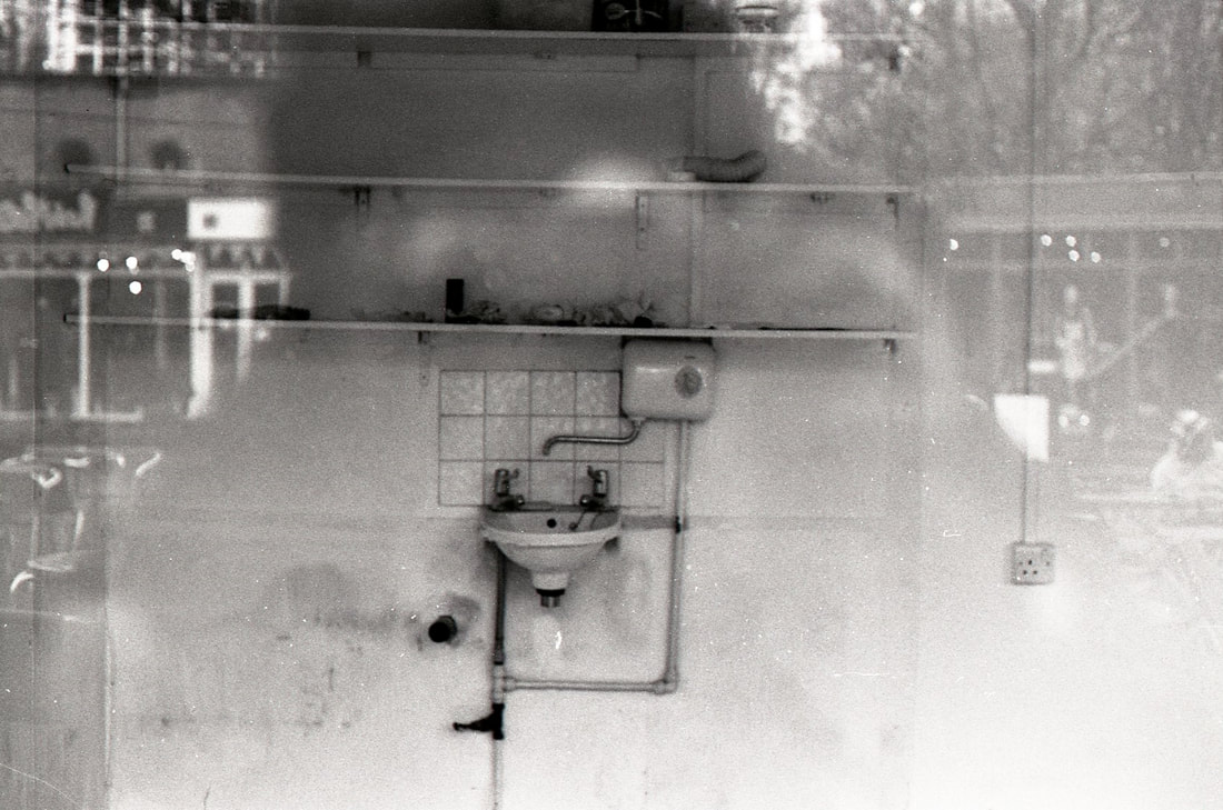 A black and white snap of a sink taking outside through the window with photographer's reflection on it. 