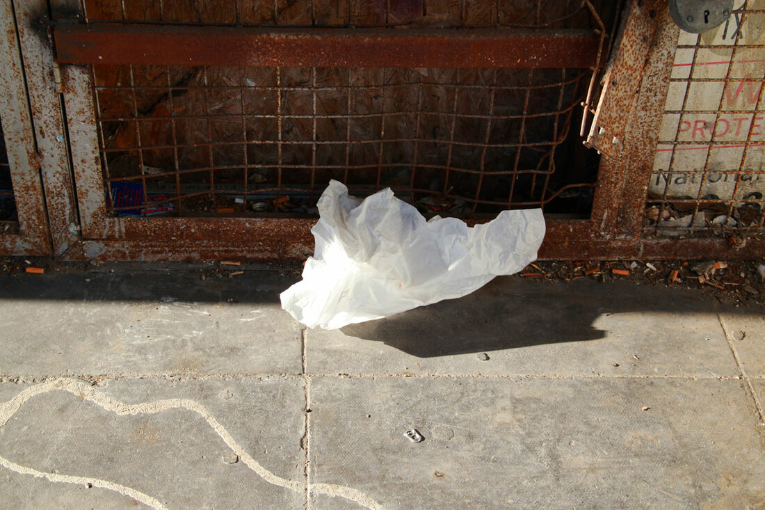 A white plastic sits on the pavement in front of a rusty fence with shadows on the floor.