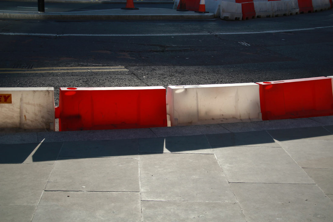 Red and white roadblocks on the side of the road create shadows on the floor by the sunshine.