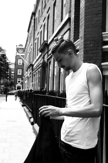 Black and white photo of a male model in white shirt standing against the fence on the street of London 